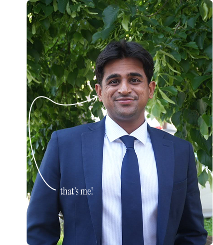 Vyomesh standing outdoors in a navy suit with a handwritten note saying that's me